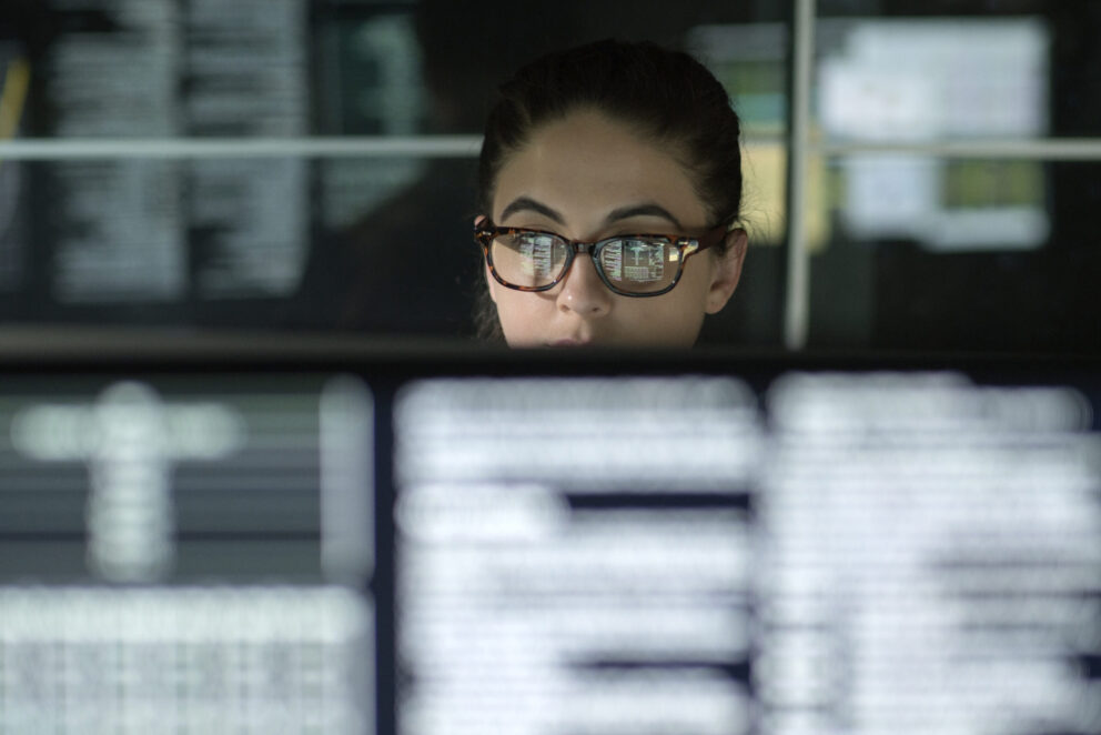 A young woman is surrounded by monitors & their reflections displaying scrolling text & data.